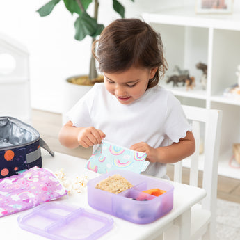A girl eats lunch with Bumkins Lunchbox Legends 6-Piece Set: Unicorns Purple Jelly near a lunch bag at a white table in a playroom.