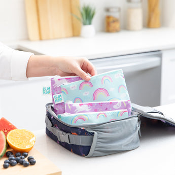 A hand places Bumkins Reusable Snack Bag 4 Pack: Unicorns and Rainbows in an open lunch bag on a kitchen counter next to fruit.