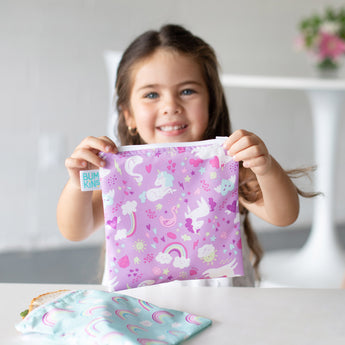 A smiling girl holds bags from Bumkins Snack In Style 3-Piece Set: Unicorns and Rainbows.