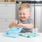 A smiling boy places pretzels into bags from Bumkins Lunchbox Legends 6-Piece Set: Ocean Blue Jelly at a white table.
