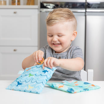 A smiling boy places pretzels into bags from Bumkins Lunchbox Legends 6-Piece Set: Ocean Blue Jelly at a white table.
