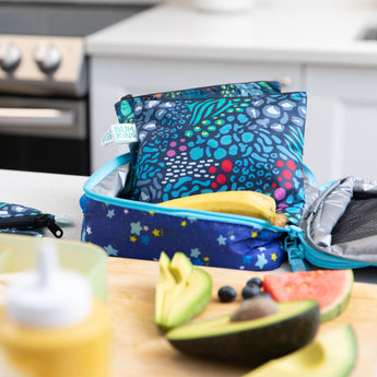 Bumkins Reusable Snack Bag, Large 2-Pack: Jungle & Animal Prints sits in an open lunch bag on a kitchen counter among fruit & veggies.