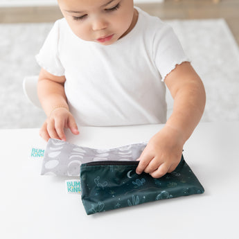 A young child in a white shirt reaches for Bumkins Reusable Snack Bag 4 Pack: Desert Night on a white table.