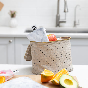 A Roamio by Bumkins Reusable Snack Bag, Small 2 Pack: Disco Boots sits in a lunch bag on a kitchen counter with fruit on a cutting board.