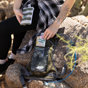 A person packs Roamio by Bumkins Reusable Snack Bag, Small 2 Pack: Motley Club in a backpack outdoors on rocks.