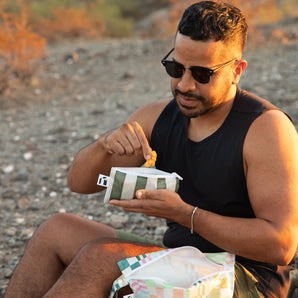 A man wearing sunglasses enjoys snacks from a Roamio by Bumkins Reusable Snack Bag, Small 2 Pack: Motley Club while sitting on rocky ground.
