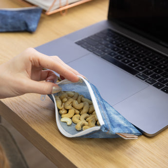 A hand opens a Roamio by Bumkins Reusable Snack Bag, Small 2 Pack: Denim on Denim next to a laptop, revealing cashews inside.
