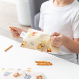 A child in a white shirt holds a Bumkins Reusable Snack Bag 4 Pack: Disney Pooh Bear + Friends with pretzel sticks on a white table.