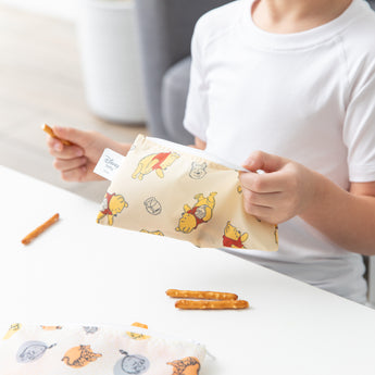 A child in a white shirt holds a Bumkins Reusable Snack Bag 4 Pack: Disney Pooh Bear + Friends with pretzel sticks on a white table.