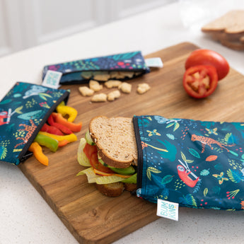 Reusable sandwich and snack bags with colorful animal and plant designs on a wooden cutting board. One bag holds a sandwich with lettuce and tomato, another contains sliced bell peppers, and a third has snacks. Sliced tomatoes are nearby.
