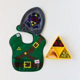 Bumkins Caped SuperBib®: The Legend of Zelda™ Link lays flat next to a yellow triangle-shaped plate with fruit on a white background.