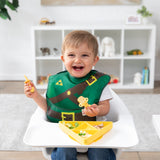 A boy wearing a Bumkins Caped SuperBib®: The Legend of Zelda™ Link smiles in a high chair, holding yellow utensils by a matching plate.
