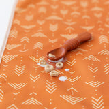 A Bumkins Silicone Dipping Spoons 3 Pack: Rocky Road rests among cereal pieces and milk splatter on an orange abstract printed mat.