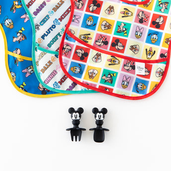 Utensils from Bumkins Grip Dish Essential Infant 3-Piece Mealtime Set: Disney Mickey Mouse rest near 3 colorful waterproof bibs.