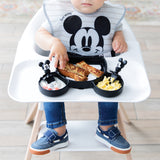 A child wearing a Mickey Mouse bib eats breakfast with Bumkins Grip Dish Essential Infant 3-Piece Mealtime Set: Disney Mickey Mouse.