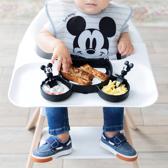 A child wearing a Mickey Mouse bib eats breakfast with Bumkins Grip Dish Essential Infant 3-Piece Mealtime Set: Disney Mickey Mouse.