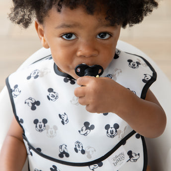 A curly-haired boy in a black & white bib chews a utensil from Bumkins Grip Dish Essential Infant 3-Piece Mealtime Set: Disney Mickey Mouse.