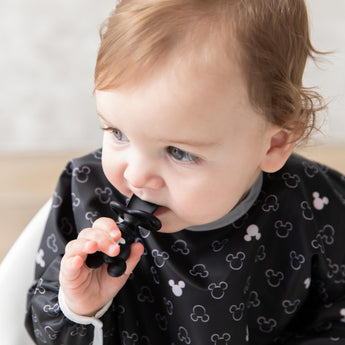 A baby in a black Mickey-themed bib chews a Bumkins Silicone Chewtensils®: Mickey Mouse.