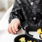Close-up of a child in a black sleeved bib eating eggs with Bumkins Grip Dish Essential Infant 3-Piece Mealtime Set: Disney Mickey Mouse.