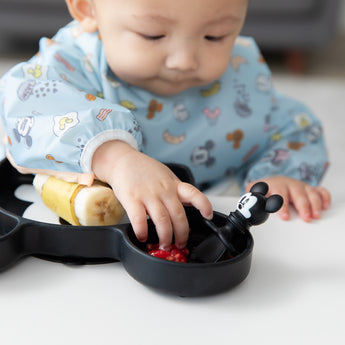 A boy in a blue bib reaches for a Bumkins Silicone Chewtensils®: Mickey Mouse sitting on a Mickey Mouse-shaped plate with fruit.