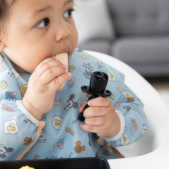 Close-up of a child chewing fruit while holding a utensil from Bumkins Grip Dish Essential Infant 3-Piece Mealtime Set: Disney Mickey Mouse.