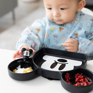 A child curiously reaches for Bumkins Grip Dish Essential Infant 3-Piece Mealtime Set: Disney Mickey Mouse with fruit and yogurt.