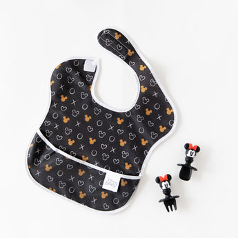 Bumkins Silicone Chewtensils®: Minnie Mouse rest next to a black & white Minnie Mouse-patterned bib on a white background.