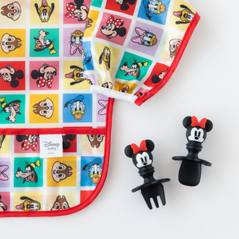 Close-up of Bumkins Silicone Chewtensils®: Minnie Mouse next to a colorful Disney character print bib on a white background.