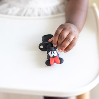 A young child in a white shirt holds a Bumkins Silicone Chewtensils®: Minnie Mouse.