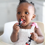 A girl in a high chair wearing a white dress chews Bumkins Silicone Chewtensils®: Minnie Mouse.