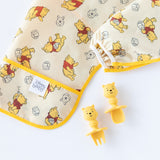 Close-up of a folded Bumkins Sleeved Bib: Winnie Loves Hunny on a white background, with Winnie the Pooh-shaped utensils nearby.