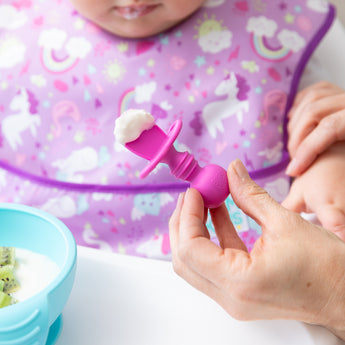 A baby with a unicorn bib eats yogurt using Bumkins Silicone Chewtensils®: Fuchsia, surrounded by silicone utensils and a kiwi bowl.