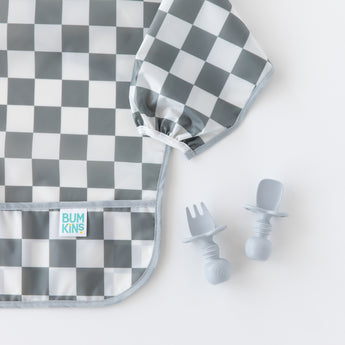Bumkins Silicone Chewtensils®: Gray rest next to a gray & white checkered sleeved bib on a white background.