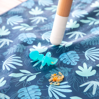 A spoon from Bumkins Grip Dish Deluxe Infant 5-Piece Mealtime Set: Blue Jelly rests atop a blue patterned floor mat next to food splatter.