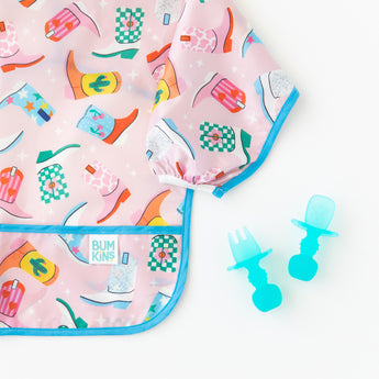 Close-up of Bumkins Sleeved Bib: Boots on a white background next to blue transparent toddler utensils.