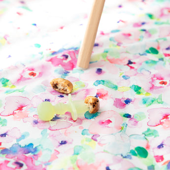 A Bumkins Silicone Chewtensils®: Green Jelly lays on top of a floral mat next to pancake pieces under a high chair leg.