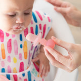 An adult hand uses a Bumkins Silicone Chewtensils®: Pink Jelly to feed a child with a yogurt-covered mouth in a colorful bib.