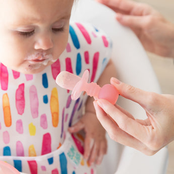 An adult hand uses a Bumkins Silicone Chewtensils®: Pink Jelly to feed a child with a yogurt-covered mouth in a colorful bib.