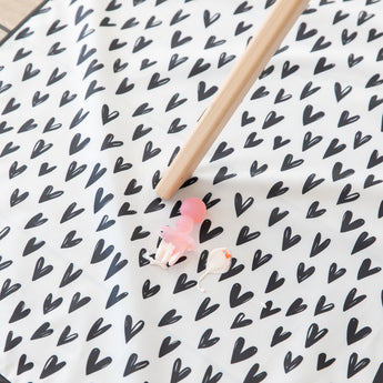 A Bumkins Silicone Chewtensils®: Pink Jelly rests on top of a black & white heart print mat with yogurt splatter under a high chair leg.