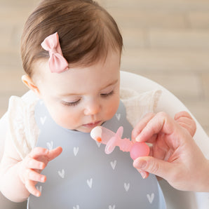 An adult hand feeds a baby in a high chair wearing a bow and a gray bib with a Bumkins Silicone Chewtensils®: Pink Jelly.