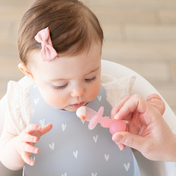 An adult hand feeds a baby in a high chair wearing a bow and a gray bib with a Bumkins Silicone Chewtensils®: Pink Jelly.
