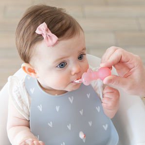 A baby with a bow in a high chair wears a Bumkins Silicone Bib 2 Pack: Hearts and Rainbows, being fed with a pink spoon by an adult hand.