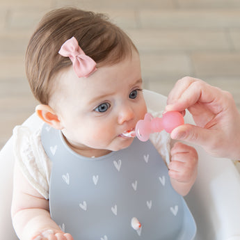 A baby in a gray Bumkins Silicone Bib 2 Pack: Hearts and Rainbows is fed by an adult hand with a pink spoon.