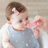 An adult hand uses a spoon from Bumkins Grip Dish Deluxe Infant 5-Piece Mealtime Set: Pink Jelly to feed a baby in a gray bib.