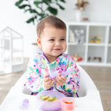 A baby grins in a high chair, holding Bumkins Silicone Chewtensils®: Purple Jelly. White shelves and a plant are visible nearby.
