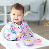 A baby in a high chair smiles, wearing a floral bib, holding Bumkins Silicone Chewtensils®: Purple Jelly, with a bowl of food nearby.
