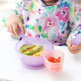 A child in a colorful bib enjoys avocado and rice from a purple bowl with Bumkins Silicone Chewtensils®: Purple Jelly, plus a cup of tomatoes.