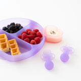 A purple plate holds waffles and berries with a heart-shaped bowl and Bumkins Silicone Chewtensils®: Purple Jelly nearby on a white background.