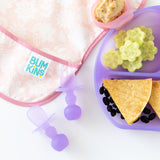 Close-up of Bumkins Silicone Chewtensils®: Purple Jelly next to a pink bib and a purple divided plate with food on a white background.