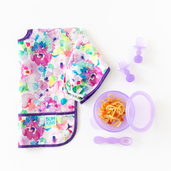A floral baby bib with Bumkins Silicone Chewtensils®: Purple Jelly, a purple bowl with spaghetti, and a spoon on a white background.